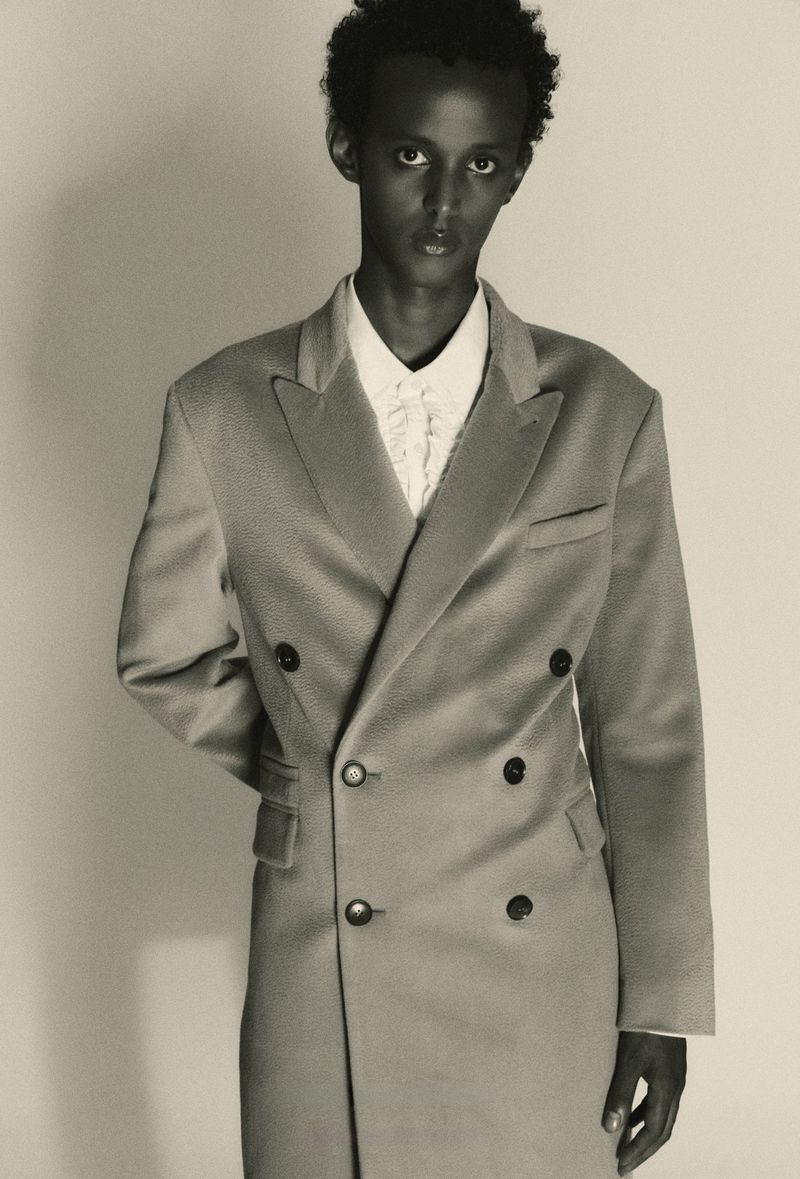 Portrait of a model wearing a double-breasted coat and white collared shirt against a plain light background.