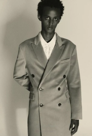 Portrait of a model wearing a double-breasted coat and white collared shirt against a plain light background.
