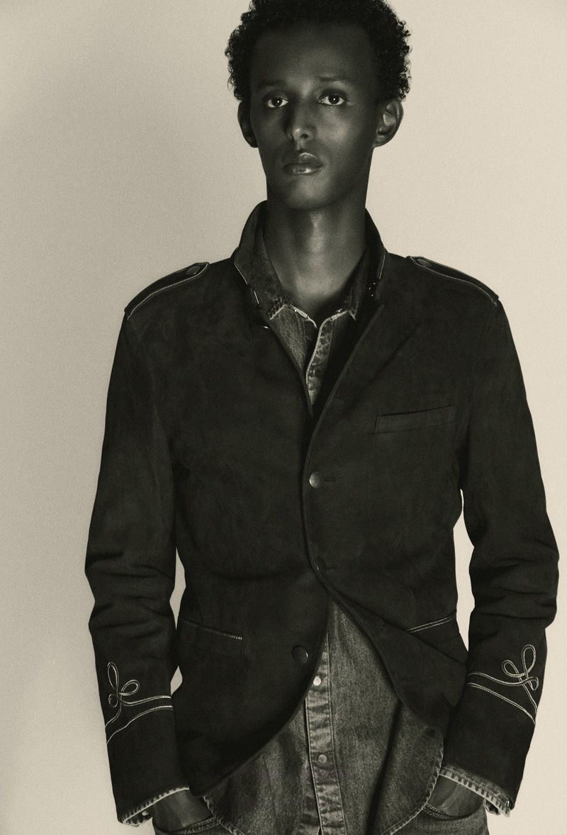 Black and white portrait of a young Black male model wearing a dark tailored jacket and denim shirt against a seamless background.