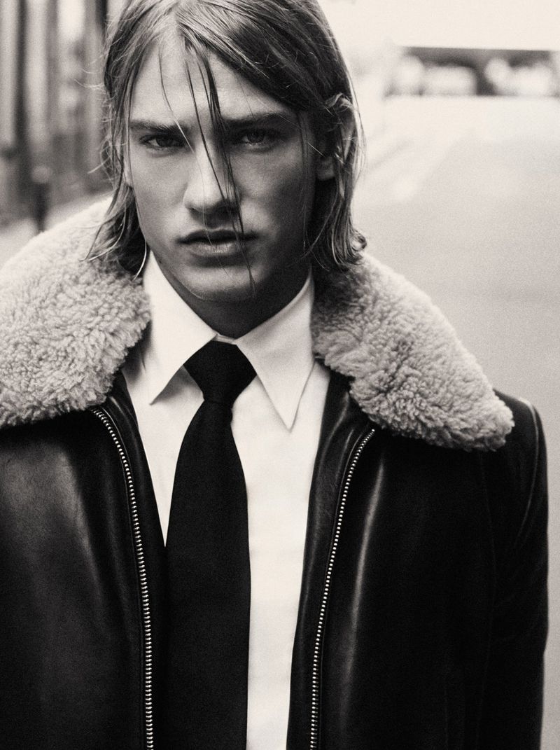 Monochrome portrait of a male model wearing a dark leather jacket with a shearling collar over a white shirt and black tie.