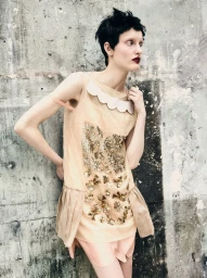 Model with short dark hair in beige embellished dress posing against a weathered concrete wall for Mixte.