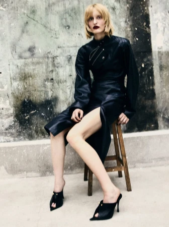 Model wearing a black leather dress seated on a wooden stool against a distressed concrete wall