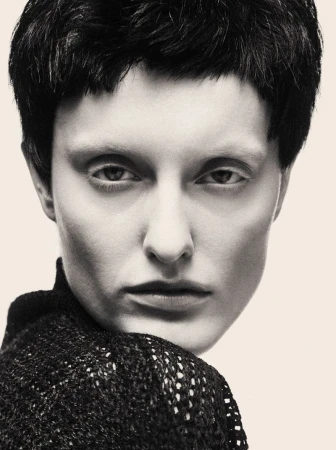 Close-up portrait of a model with short dark hair wearing a textured black turtleneck against a neutral background.