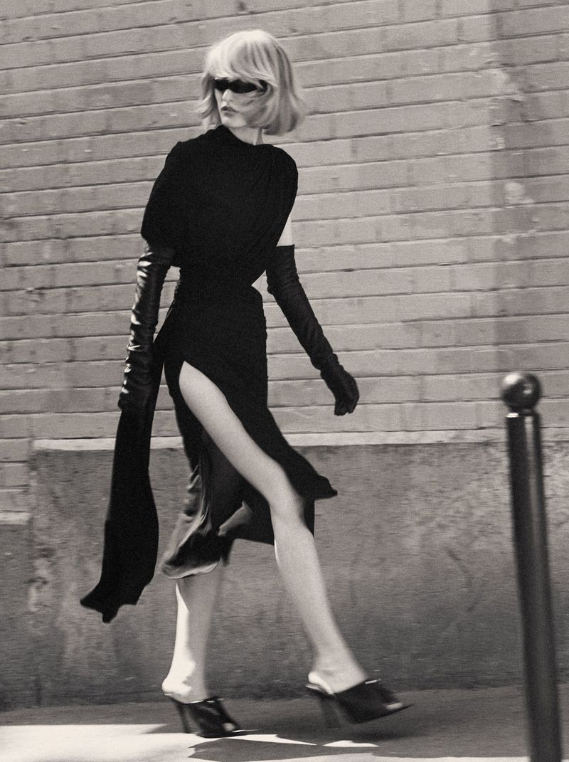 Model in a black dress with long gloves walking outdoors past a brick wall for Mixte Perfect Strangers