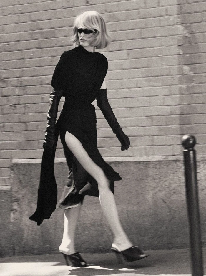 Model in a black dress with long gloves walking outdoors past a brick wall for Mixte Perfect Strangers