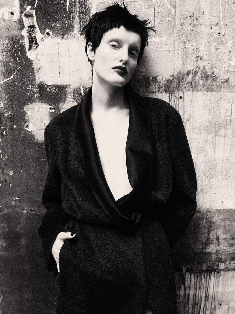 Model with short dark hair in a dark coat posing against a textured concrete wall for Mixte Perfect Strangers