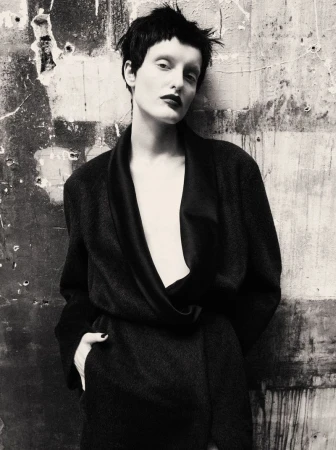 Model with short dark hair in a dark coat posing against a textured concrete wall for Mixte Perfect Strangers