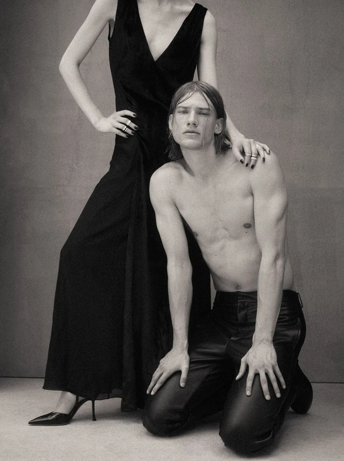 Monochrome portrait of a woman in a black dress standing behind a shirtless man kneeling in leather trousers.