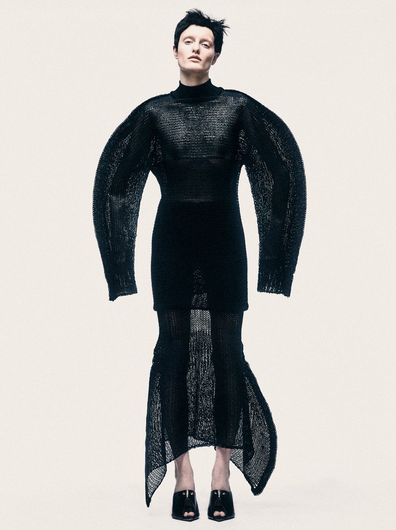 Model standing against a plain background wearing a black knit sweater with exaggerated long sleeves