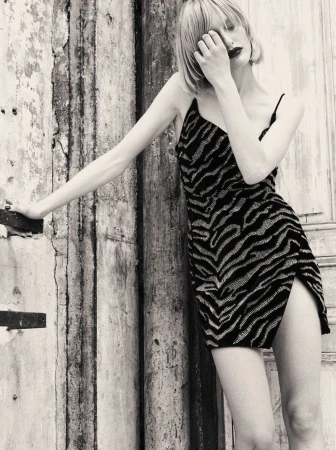 Monochrome fashion editorial portrait of a woman in a patterned slip dress leaning against a weathered wooden wall.
