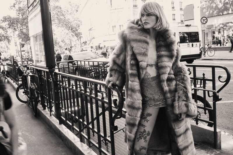 Model wearing a large fur coat standing by a city street railing in black and white