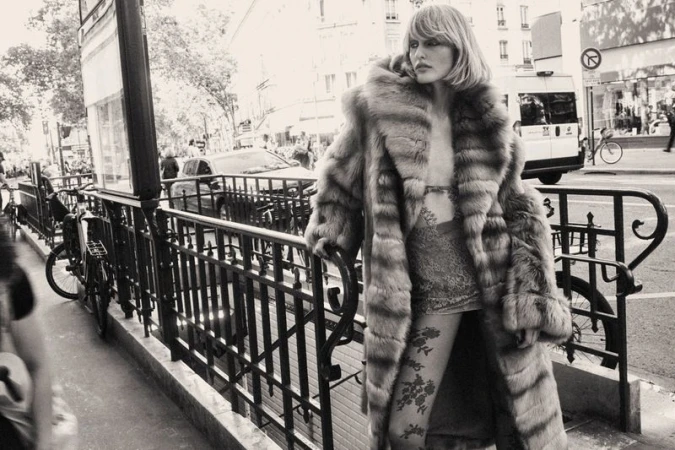 Model wearing a large fur coat standing by a city street railing in black and white