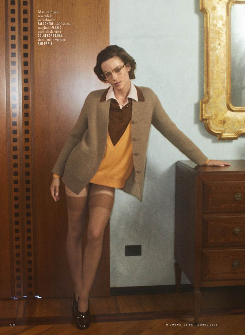 Model wearing a camel cardigan, yellow vest, and stockings leaning against a wood-paneled wall indoors.
