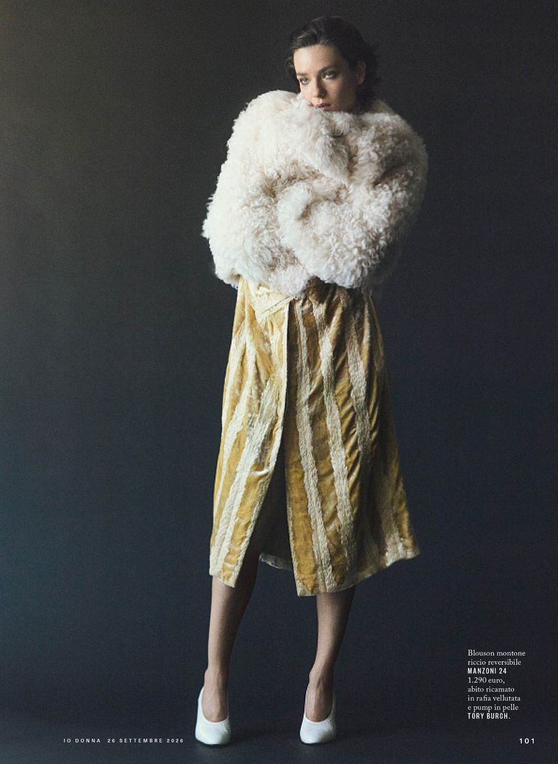 Model in a fluffy white textured coat and gold patterned midi dress standing against a dark grey background.