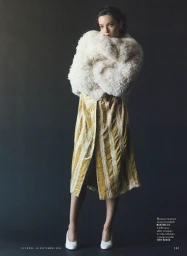Model in a fluffy white textured coat and gold patterned midi dress standing against a dark grey background.