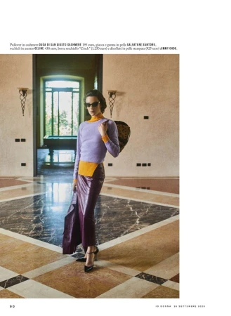 Model wearing a lavender sweater and dark skirt standing in an expansive room with marble flooring and large doorways.