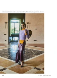 Model wearing a lavender sweater and dark skirt standing in an expansive room with marble flooring and large doorways.