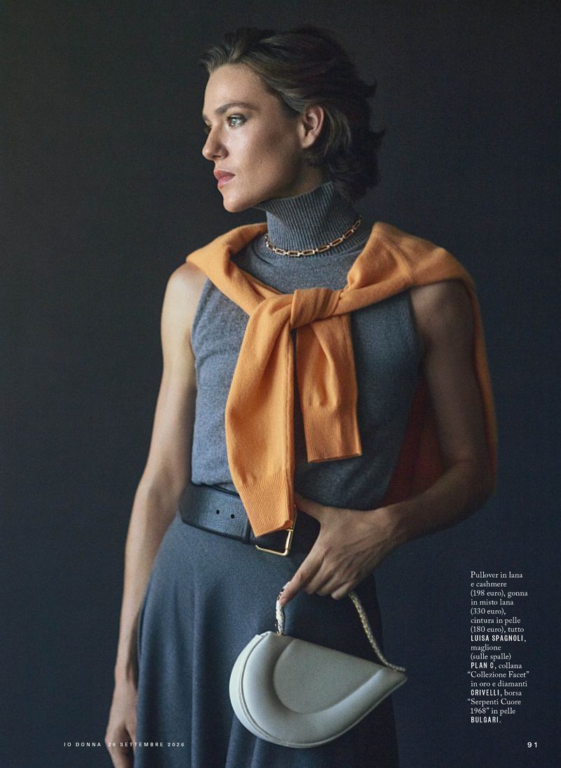 Model wearing grey turtleneck sweater with orange jumper tied over shoulders, holding small white handbag.