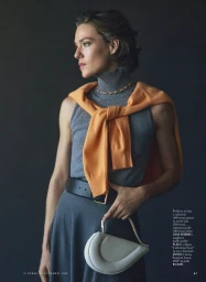 Model wearing grey turtleneck sweater with orange jumper tied over shoulders, holding small white handbag.