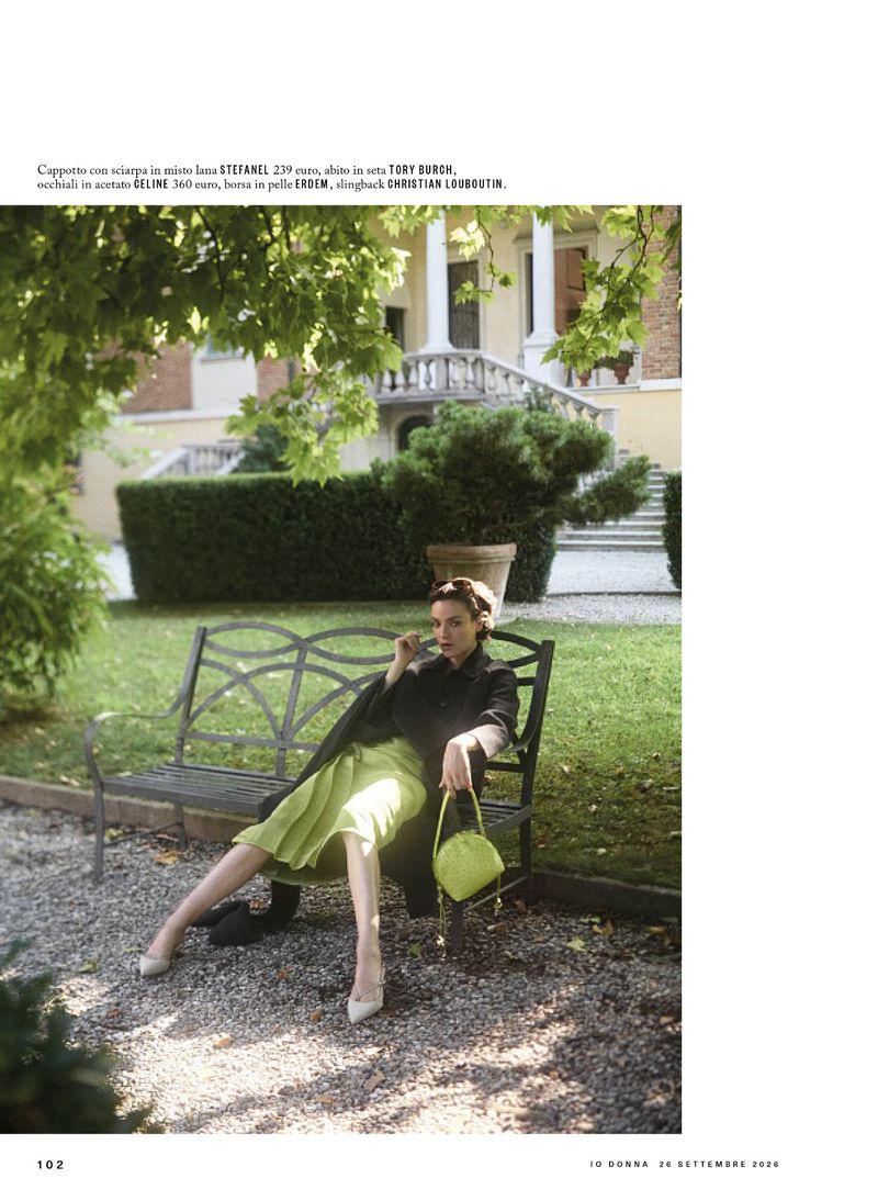 Model seated on a garden bench in front of a historic building, wearing a dark coat and holding a handbag.