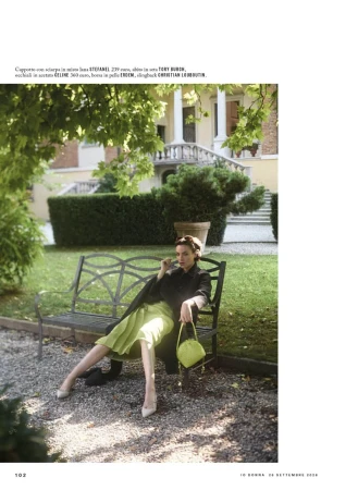 Model seated on a garden bench in front of a historic building, wearing a dark coat and holding a handbag.