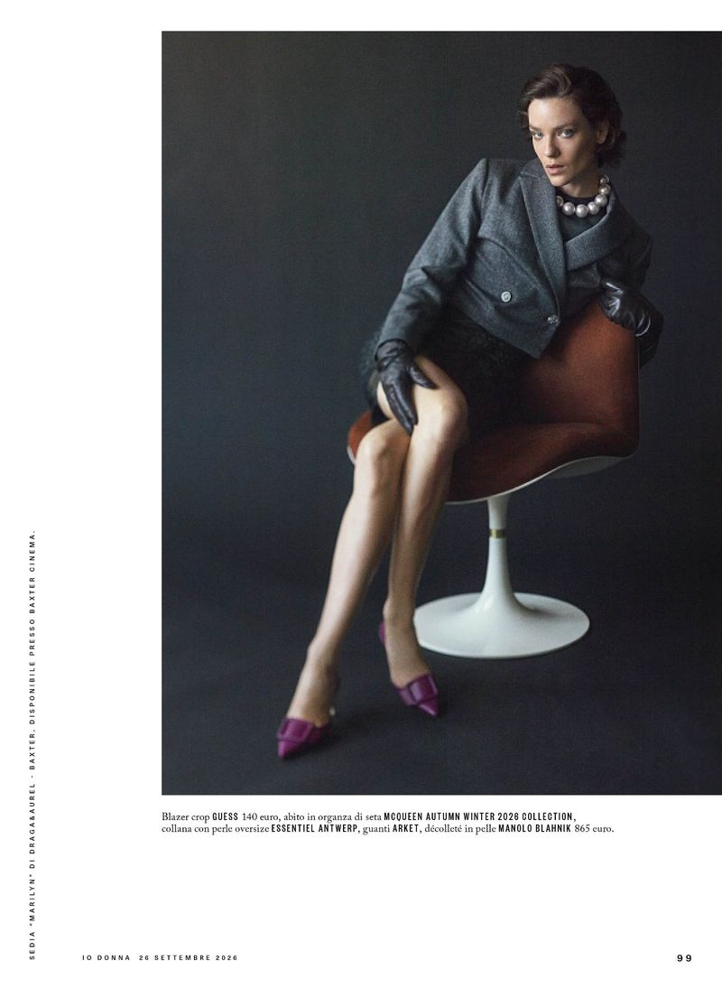 Model seated on a modern chair wearing a grey blazer, black gloves, pearl necklace, and purple heels against a dark backdrop.