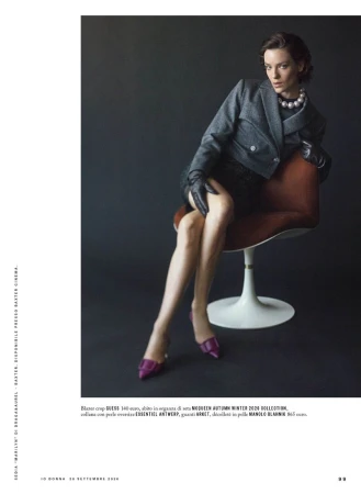 Model seated on a modern chair wearing a grey blazer, black gloves, pearl necklace, and purple heels against a dark backdrop.