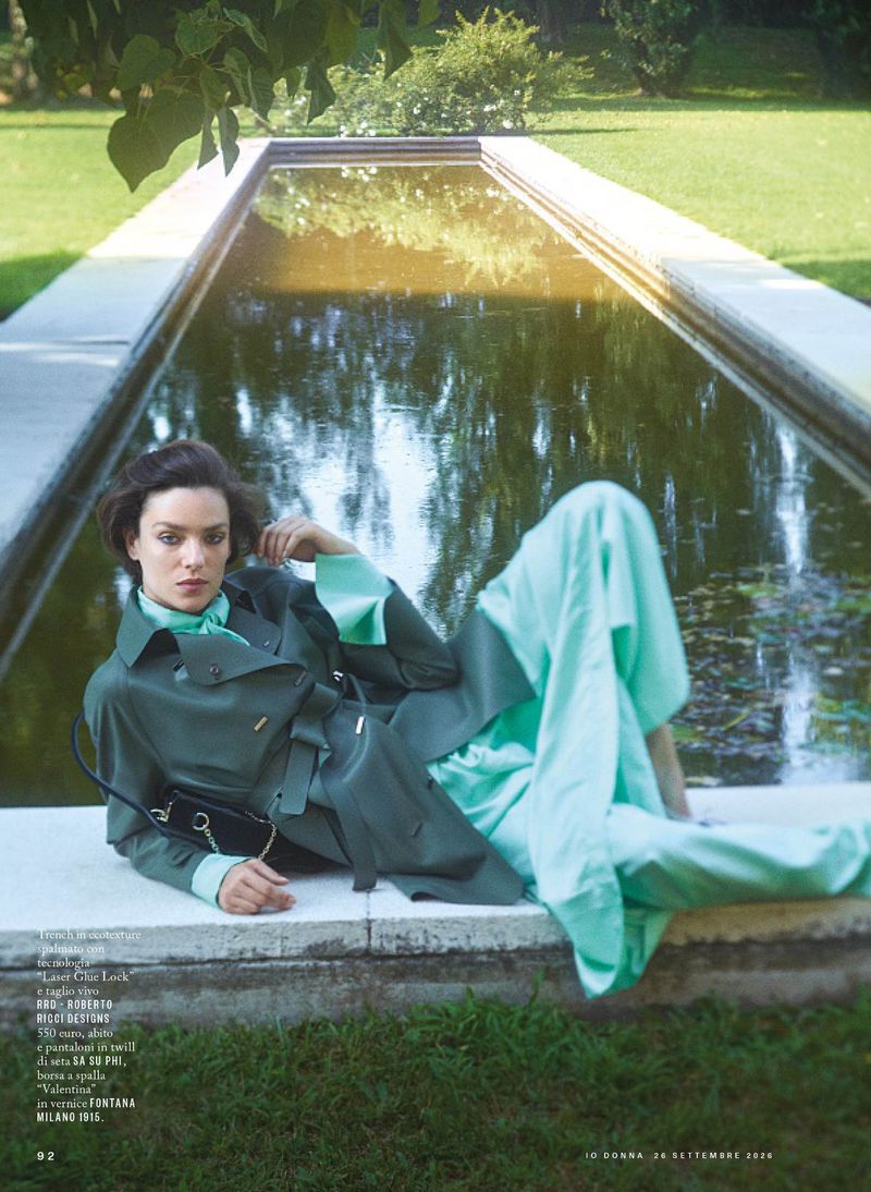 Model lying beside a reflecting pool wearing a green coat and light blue trousers for iO Donna editorial