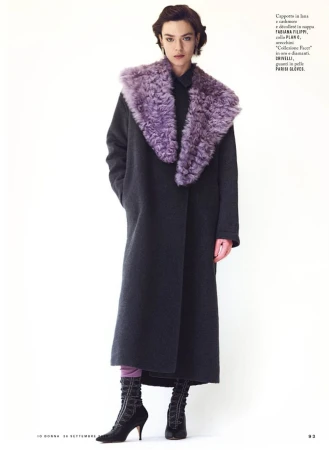 Model wearing a dark long coat with a prominent purple fur collar against a plain white background