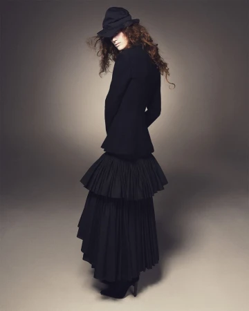 Model in black layered skirt and blazer wearing a dark hat against a grey studio backdrop for D Repubblica