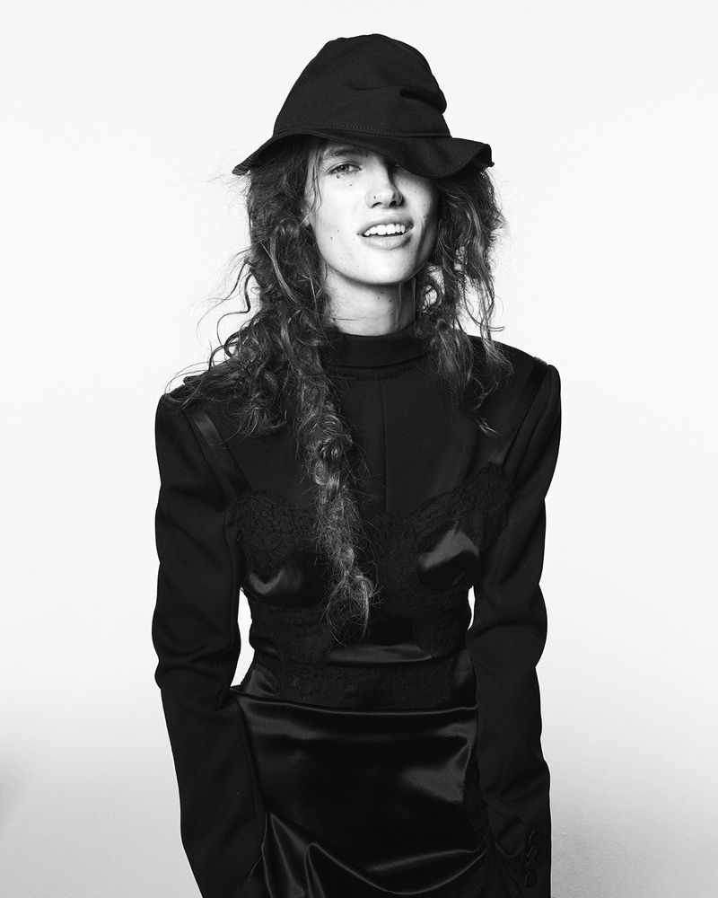 Model wearing a black hat and black outfit with long curly hair against a plain white background