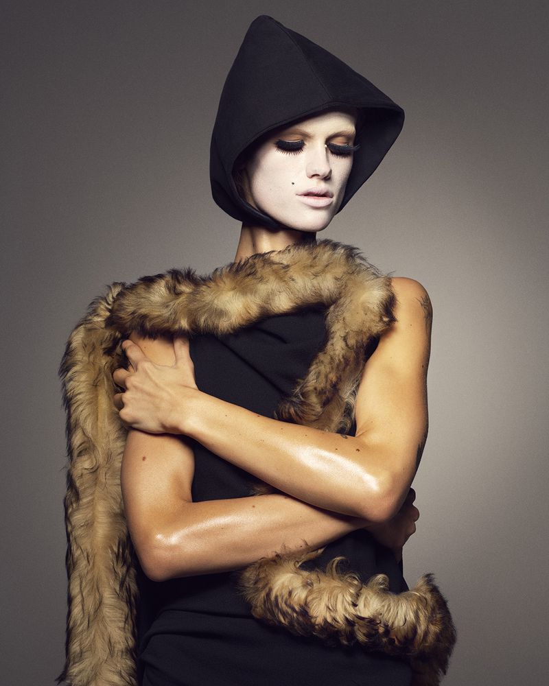Model wearing a dark fitted hood and draped fur scarf against a grey background