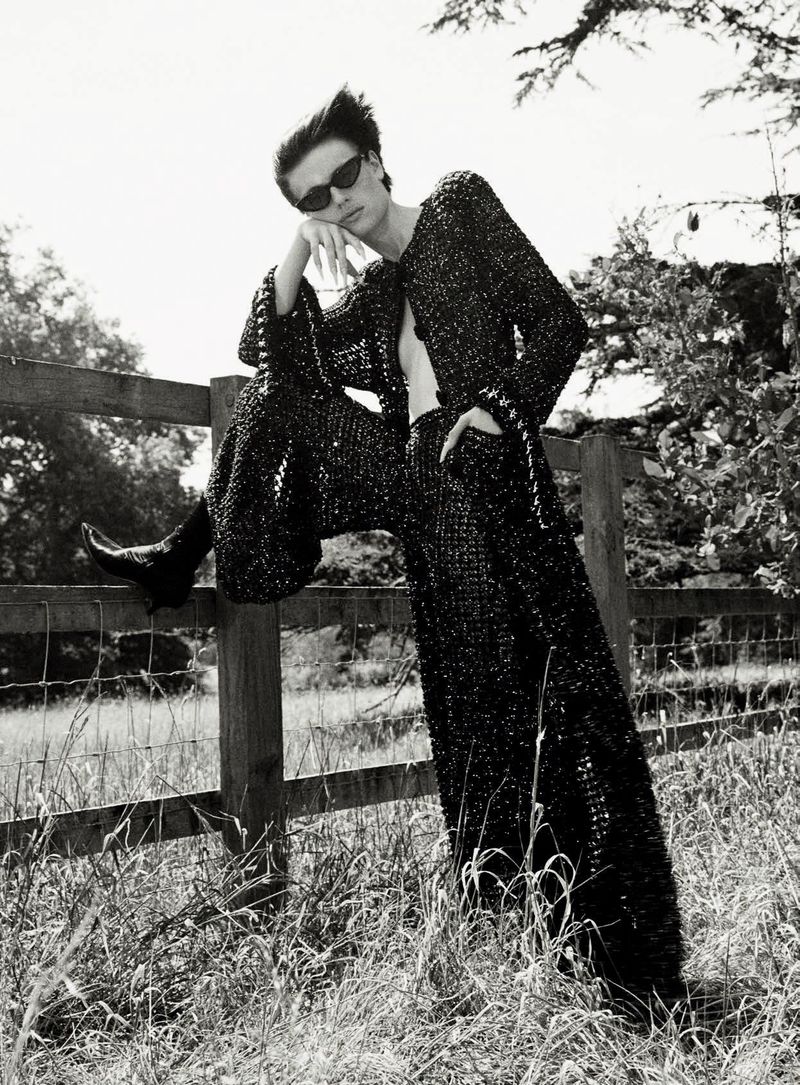 Model in dark sunglasses and textured black outfit leaning on a wooden fence outdoors.