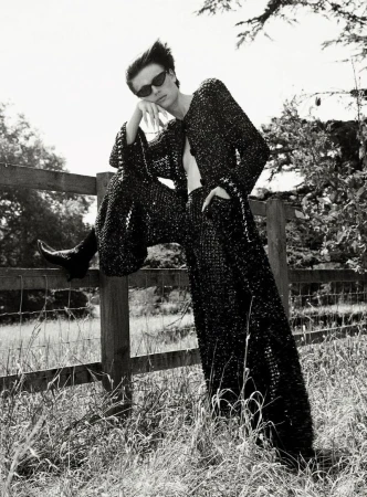 Model in dark sunglasses and textured black outfit leaning on a wooden fence outdoors.