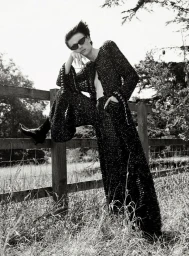 Model in dark sunglasses and textured black outfit leaning on a wooden fence outdoors.