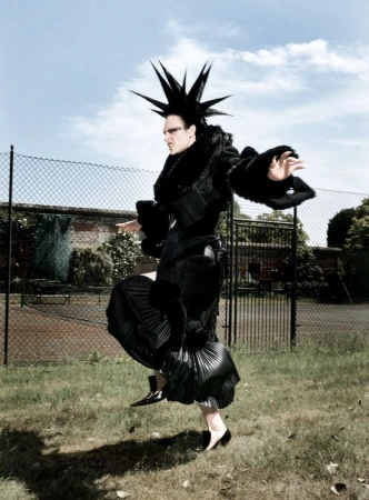 Model with spiky black hair in a black coat and pleated skirt posing outdoors against a fence