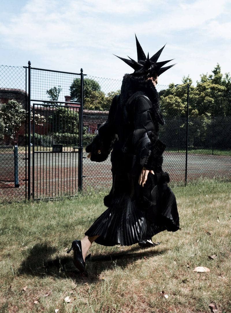 Model in a black avant-garde spiky headpiece and black pleated skirt walking past a chain-link fence on a grassy field