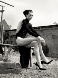 Monochrome fashion portrait of a model in a black dress seated on a wooden bench outdoors against a brick wall.