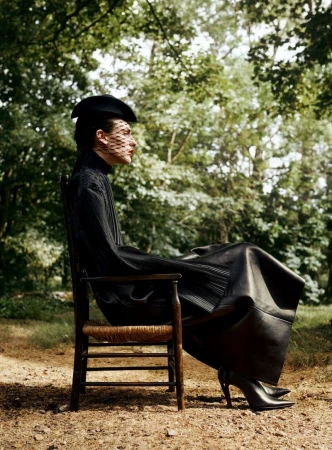 Model seated on a wooden chair in a forest wearing a black dress and netted hat for D Repubblica photographed by Ned Rogers