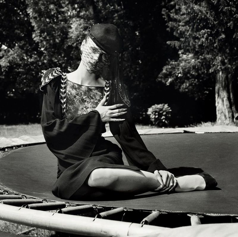 Model seated on a trampoline outdoors wearing a dark dress with embellished shoulders and a veiled cap in a black and white photograph.