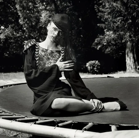 Model seated on a trampoline outdoors wearing a dark dress with embellished shoulders and a veiled cap in a black and white photograph.