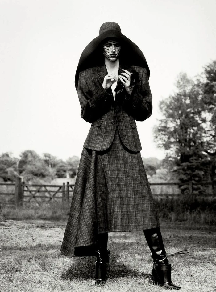 Model in plaid skirt suit and oversized wide-brim hat standing outdoors for D Repubblica