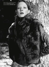 Model leaning against a large tree trunk wearing a dark fur vest and collared shirt in a monochrome outdoor editorial.