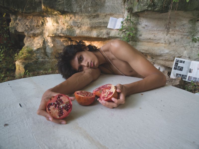 Shirtless young man resting on a white-covered surface, holding halved pomegranates against a rocky stone wall.