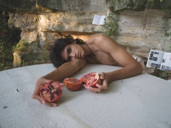 Shirtless young man resting on a white-covered surface, holding halved pomegranates against a rocky stone wall.