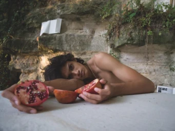 Young man lying on a stone surface holding halves of a split pomegranate against a rocky backdrop.