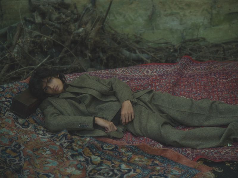Model wearing an olive green suit lying on a patterned red rug outdoors with a pile of branches behind them.