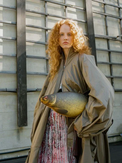 Red-haired female model holding a fish-shaped handbag wearing an oversized taupe coat and patterned skirt