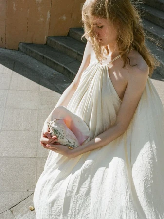 Model in white halter dress holding a large conch shell while seated near outdoor concrete stone stairs.