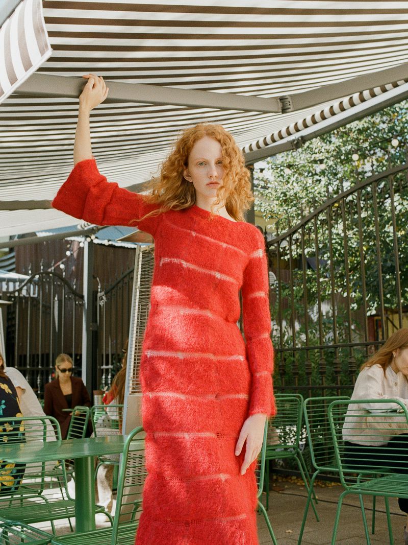Red-haired female model in a textured red dress standing under a striped awning outdoors for Cake Magazine